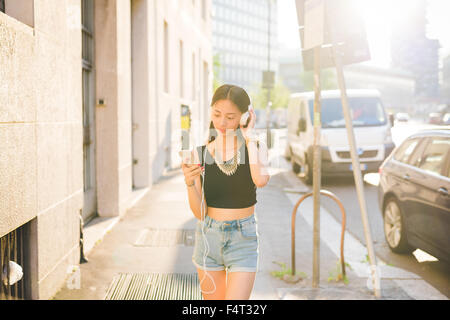Knie-Figur des jungen schönen asiatischen Frau Wandern im Freien in der Stadt, hören Musik mit Kopfhörer, während mit einem Smartphone auf der Suche nach unten die Bildschirm - Technik, Musik, entspannende Konzept Stockfoto