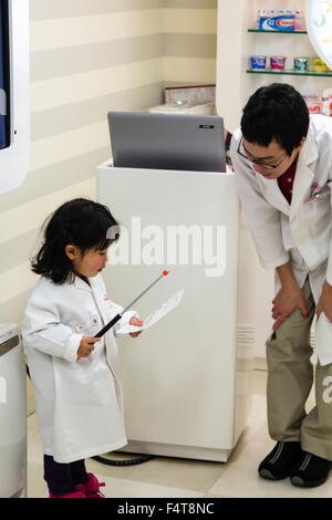 Japan, Nishinomiya, KidZania, Rollenspiele, Freizeitzentrum für Kinder. Kind, Mädchen Wissenschaftler, im weißen Mantel, Vortrag, erwachsenen Mann beobachten. Stockfoto