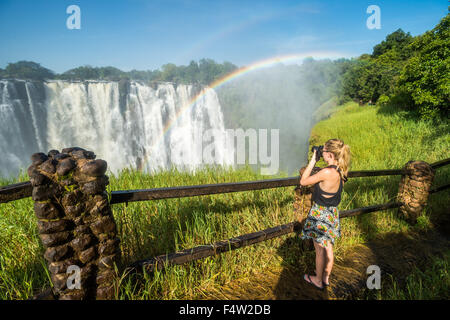 LIVINGSTONE, Sambia - Frau Fotografieren von Victoria Falls Wasserfall mit Regenbogen Stockfoto