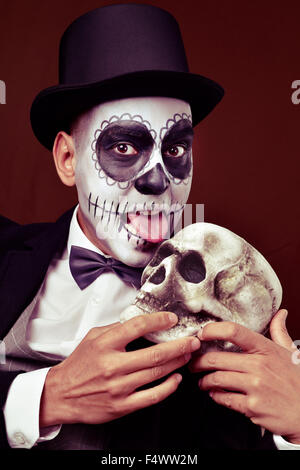 ein junger Mann mit mexikanischen Calaveras Make-up tragen, Zylinder und Fliege, leckt einen gruseligen Schädel Stockfoto