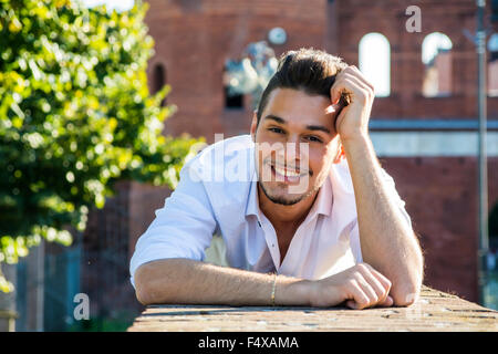 Attraktive junge Mann Verlegung auf Mauer, Blick in die Kamera Lächeln an einem sonnigen Tag Stockfoto