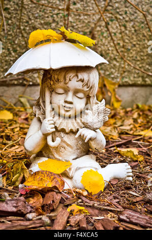 Eine verwitterte Statue eines jungen Mädchens mit einem Regenschirm ein ein kleinen Vogel, umgeben von trockene Blätter und Rinde Chips in meinem Hinterhof. Stockfoto