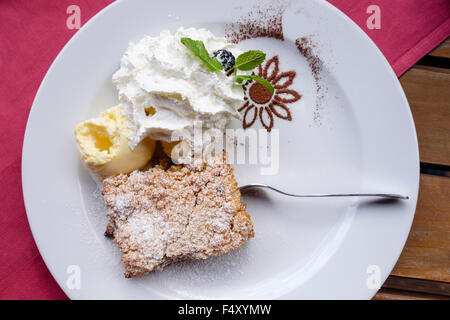 Weißer Teller von Apple crumble Dessert mit Eis und Schokoladenpulver mit einer Gabel auf einem Holztisch mit einer roten Tischdecke Stockfoto