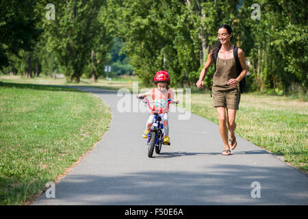 Ein drei Jahre altes Mädchen, trug einen Roten Helm, ist Radfahren erstmals ihre Mutter läuft neben ihr Stockfoto