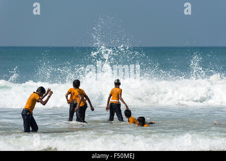 Eine Gruppe von Schuljungen, tragen gelbe Hemden, steht am Strand, einige spielen auf den Wellen Stockfoto