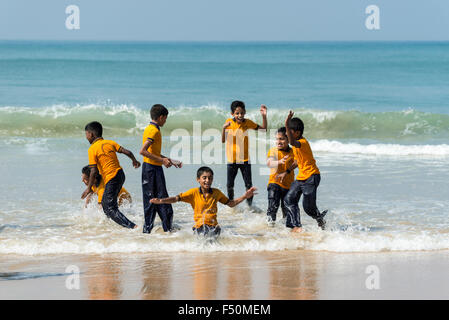 Eine Gruppe von Schuljungen, tragen gelbe Hemden, steht am Strand, einige spielen auf den Wellen Stockfoto