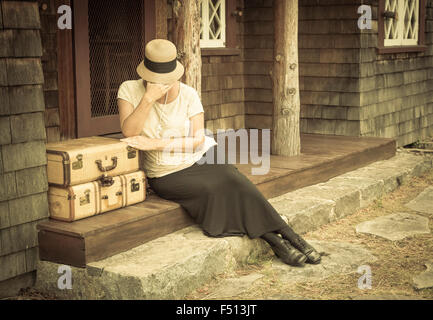 Distressed 1920er Jahre gekleideten Mädchen neben Koffer auf Veranda mit Vintage-Effekt hinzugefügt. Stockfoto