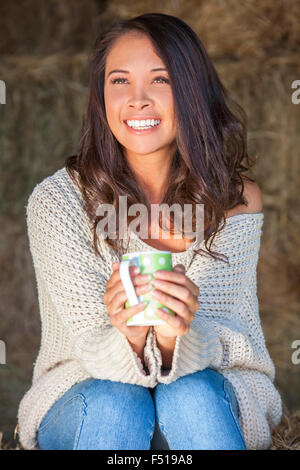Schöne glückliche asiatischen eurasischen junge Frau oder Mädchen lächelnd sitzen in der Sonne auf einem Heuballen in der Scheune, trinken Kaffee oder Tee Stockfoto