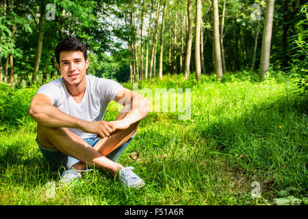 Attraktiv, Fit junger Mann entspannend sitzen auf dem Rasen Rasen auf dem Lande zwischen Bäumen und Blick in die Kamera, Lächeln Stockfoto