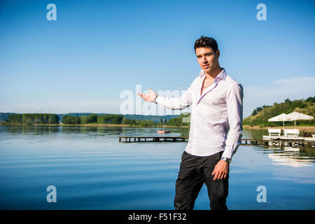 Hübscher junger Mann am Ufer eines Sees in einem sonnigen, ruhigen Tag, stehend und lädt Betrachter, den Ort zu genießen Stockfoto