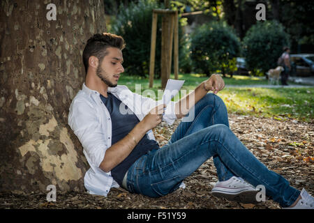 Lächelnden schönen jungen Mann, lesen oder studieren auf Papierbögen, entspannend auf grasbewachsenen Boden im Park, an Baum gelehnt Stockfoto