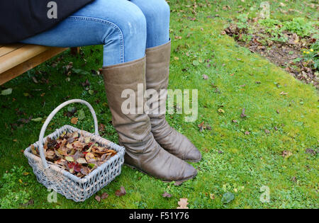 Frauen tragen Winter Gummistiefel sitzt auf einer Holzbank Garten mit einem Korb von Herbstlaub zu ihren Füßen Stockfoto