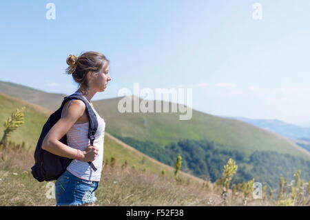 Junge, attraktive Mädchen mit einem Rucksack auf dem Rücken, auf dem Berg stehen. Grüne Wiesen und majestätische Berge in den backgr Stockfoto