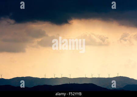 Wakayama, Japan, Windenergieanlagen im Sonnenlicht auf der Oberseite des Berges gegen gelb Himmel silhouetted, oben, sehr dunklen regen Sturm Regen Wolken. Stockfoto