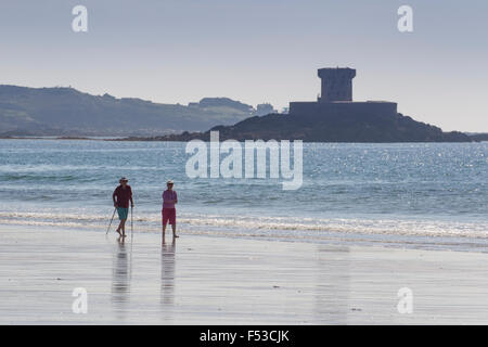 Rentner-paar zu Fuß entlang der Uferlinie auf St St Ouen Strand mit La Rocco Tower im Hintergrund Jersey Channel Islands Stockfoto