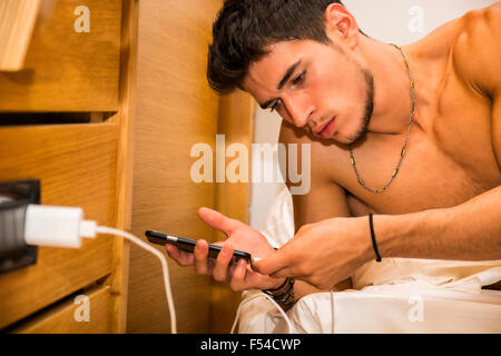 Ernsthafte wunderschöne junge Mann Connect sein Handy an ein Ladegerät während auf der Seite liegend auf dem Bett. Stockfoto