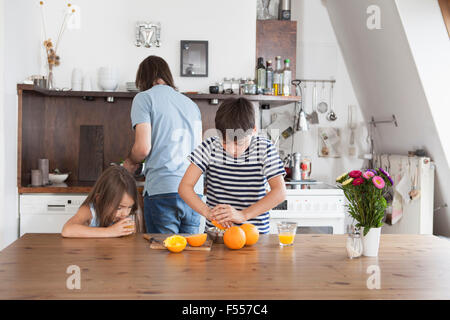 Junge drückte Orangen während Schwester trinken Saft mit Vater im Hintergrund in Küche Stockfoto