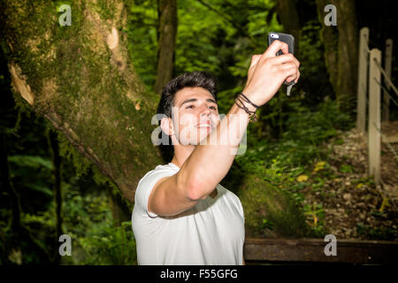 Taille bis der junge attraktive Lächeln Mann unter Selfie Selbstbildnis mit Handy im Freien im Wald Stockfoto