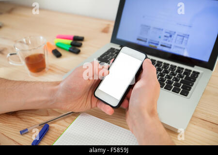 Mann mit Smartphone mit weißen Bildschirm in der Nähe von Laptop im Wohngebäude Stockfoto