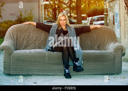 trendige blonde Frau sitzt auf einem alten Sofa in der Straße Stockfoto