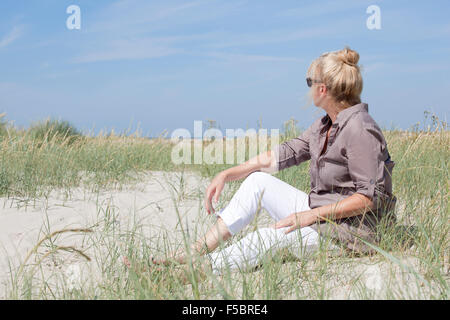 Ein Senior Frau sitzt in den Dünen auf einem Strand und schaut in die Ferne Stockfoto