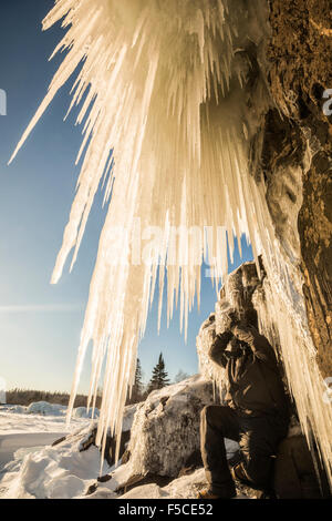 Niedrigen Seitenwinkel POV des riesigen Eiszapfen hängen von einer felsigen Klippe mit einer Person im Winter Kleidung kniend unter Eiszapfen Stockfoto