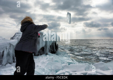 Blonde Frau kaukasischen in ihren 40ern trägt einen Wintermantel und rosa Schal wirft ein großes Stück Eis ins Wasser vom Ufer Stockfoto