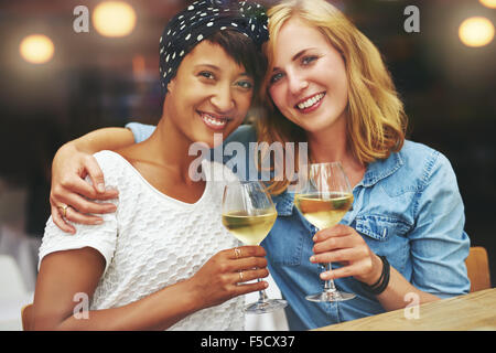 Zwei wunderschöne junge multiethnischen Frauen bei einem Glas Wein ihre Gläser in einem Toast auf die Kamera zu erhöhen, wie sie Arm, in sitzen Stockfoto