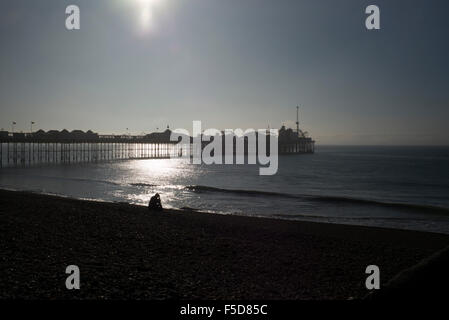 Einsame Figur sitzt am Strand vor Palace Pier von Brighton, am frühen Morgen. Stockfoto