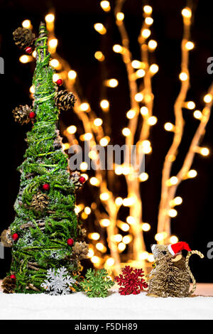 Ein Baby Maus Weihnachtsmütze mit Schneeflocken sitzt neben dem Weihnachtsbaum auf einem Nacht-Bokeh-Hintergrund. Stockfoto