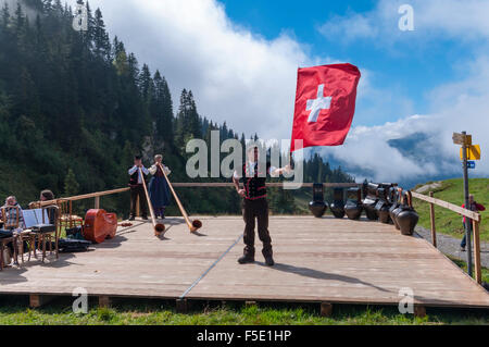 Junger Mann im Berner Tracht geben eine Fahne, die Leistung in den Schweizer Alpen zu werfen. In den Hintergrund Alphörner und Kuh-Glocken. Stockfoto