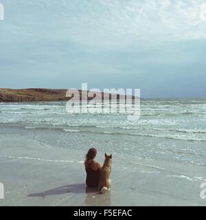 Frau sitzt neben ihrem Hund am Strand Stockfoto