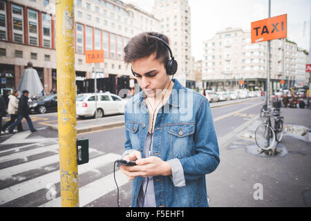 Halbe Länge des jungen schönen dunklen Alternativmodell Mann in der Stadt Musikhören mit Kopfhörer und Smartphone, auf der Suche nach unten - Musik, Technologie, entspannende Konzept Stockfoto