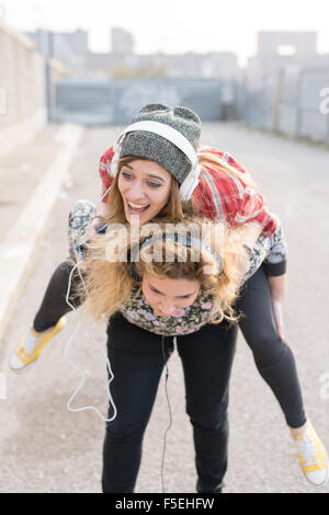 Knie-Abbildung von zwei junge lockiges und glattes blondes Haar kaukasischen Frau Musik hören mit Kopfhörern, hält die andere auf Huckepack Fahrt - Musik, Spaß, Freundschaft-Konzept Stockfoto