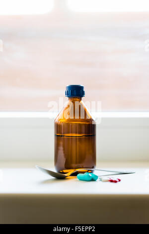 Eine Flasche Hustensaft mit einem Löffel und einige Pillen in der Nähe des Fensters Stockfoto