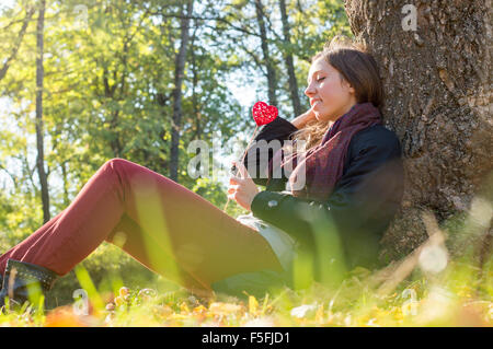 Schöne Mädchen halten rotes Herz auf einem Stick beim Sitzen auf dem Rasen. Entspannung an einem Herbsttag Stockfoto