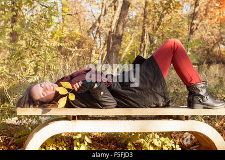 Schönes Mädchen auf der Bank liegen und halten im Herbst Blatt in einem Park in herbstlichen Farben gefärbt Stockfoto