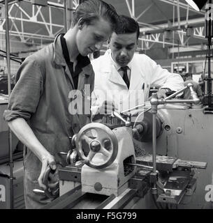 1950 historisch, ein junger männlicher Lehrling mit einem industriellen Metallschneidwerkzeug oder Drehmaschine unter der engen Aufsicht eines weiß-beschichteten Vorgesetzten. Stockfoto