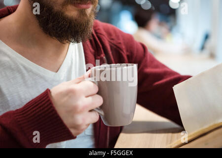 Junger Mann mit Bart hält eine Tasse Heißgetränk und Lesung Stockfoto