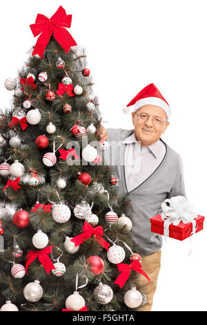 Vertikale Studioaufnahme von einem senior Mann mit Weihnachtsmütze hinter einem Weihnachtsbaum stehen und halten ein Geschenk in rotem Papier eingewickelt Stockfoto