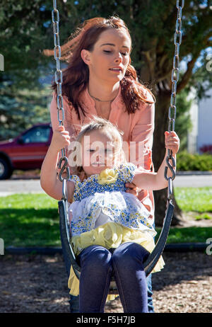 Schöne Mutter schob niedliche junge Tochter auf Schaukeln, Stadtpark Stockfoto