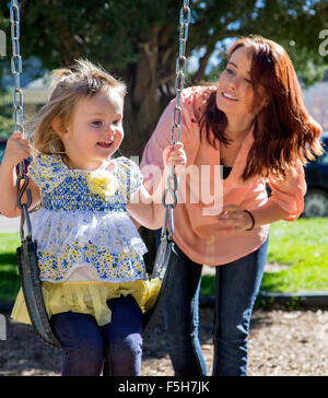 Schöne Mutter schob niedliche junge Tochter auf Schaukeln, Stadtpark Stockfoto