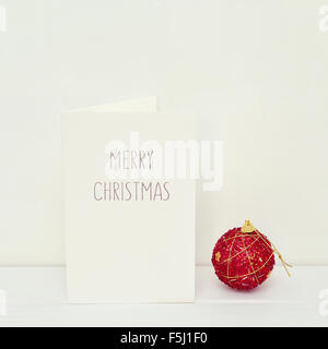 eine rote Weihnachtskugel und eine weiße Grußkarte mit dem Text Frohe Weihnachten in einer weißen Szene Stockfoto