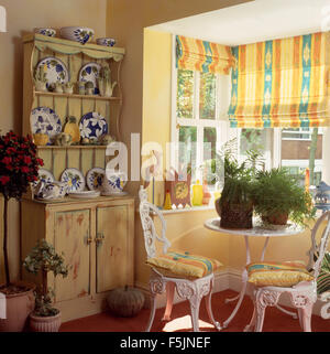 Gelbe Vorhänge am Fenster in einer neunziger Jahre Esszimmer mit weißen Schmiedeeisen Gartenstühle und Tisch und eine kleine Kiefer Kommode Stockfoto