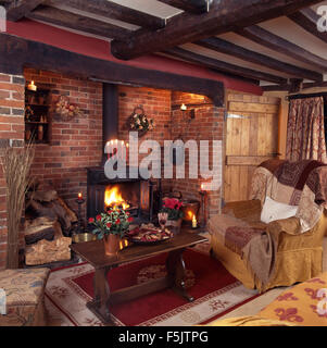 Ferienhaus Wohnzimmer Couchtisch neben Sessel vor einem hölzernen brennenden Ofen in einem Backstein Inglenook Kamin Stockfoto