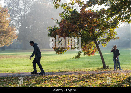 Ein Junge Skateboarder schaut auf eine ältere Roller Blader an einem sonnigen Herbsttag in einem Londoner park Stockfoto