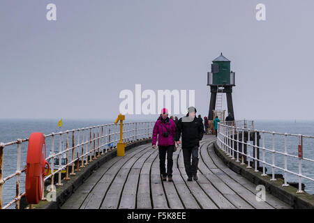 Menschen in warme Kleidung, zu Fuß auf Whitbys West Pier Erweiterung, England, UK - graue Holzbretter, Meer und Himmel, reflektieren kalten windigen Herbstwetter. Stockfoto