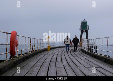 Ein Paar, das am Whitby's West Pier, England, Großbritannien, spazieren geht - graue Töne von Holzdeckenbrettern, Meer und Himmel bei kaltem, windigem, trüben Herbstwetter an der Küste. Stockfoto