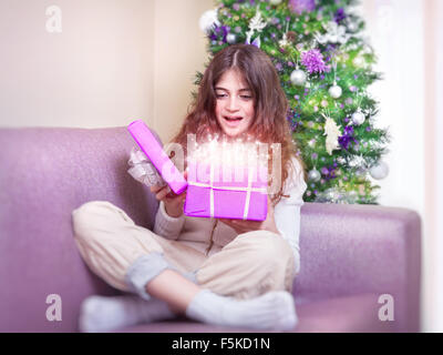 Fröhliche Teengirl öffnen Geschenkbox mit magischen glühenden vorhanden, sitzen auf der Couch in der Nähe von Weihnachtsbaum, glücklich Winterurlaub Stockfoto