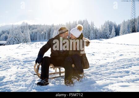 Älteres Paar auf verschneite Landschaft sitzt auf Schlitten von Angesicht zu Angesicht lächelnd, Sattelbergalm, Tirol, Österreich Stockfoto